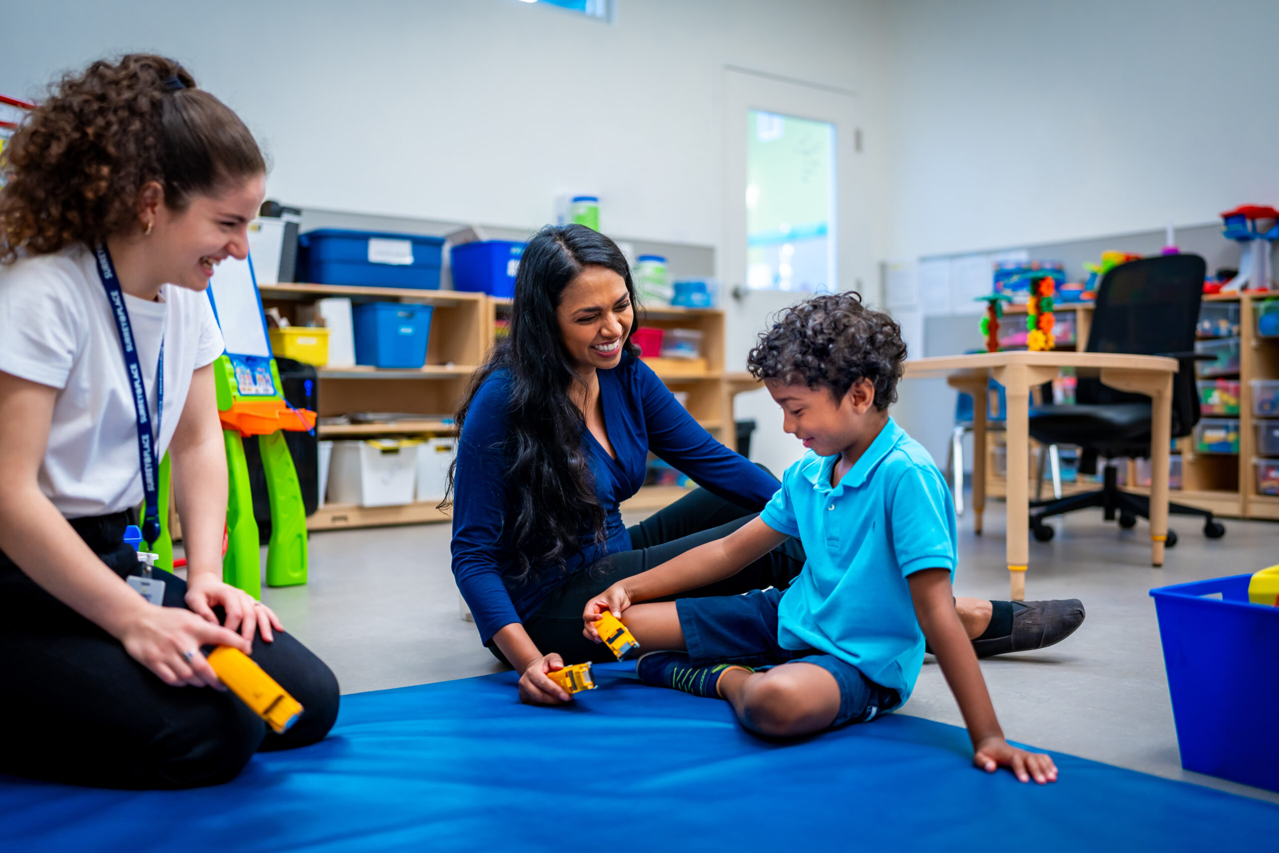 A classroom setting with three individuals sitting on a blue mat. Two adults—  
one in a white shirt and black pants, the other in a dark blue top and black pants—are   
engaging with a child wearing a light blue polo shirt and dark shorts. The child is   
holding yellow toy blocks. Behind them are shelves filled with educational materials and   
toys, and a desk with a chair.]